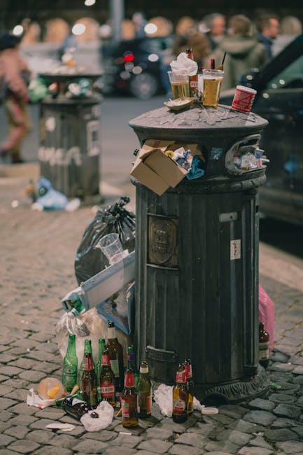 A residential area with a mound of mixed waste and rubbish placed on the grass in the foreground, including black plastic bags, cardboard boxes, and loose debris. Behind the waste, there are small, si