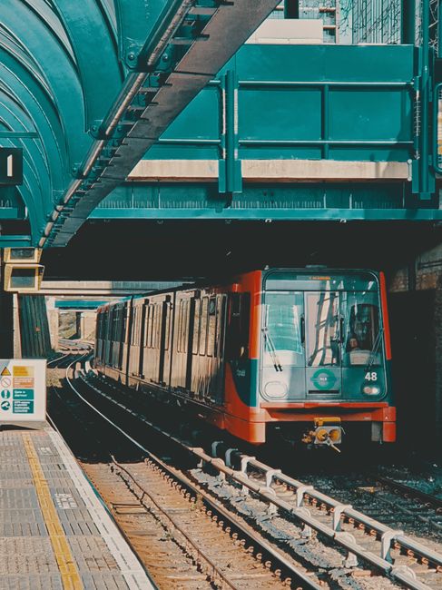 A worker standing on a train station platform next to a large handcart loaded with various bags and bulk waste materials, including black, orange, and grey rubbish bags, some with visible waste protru